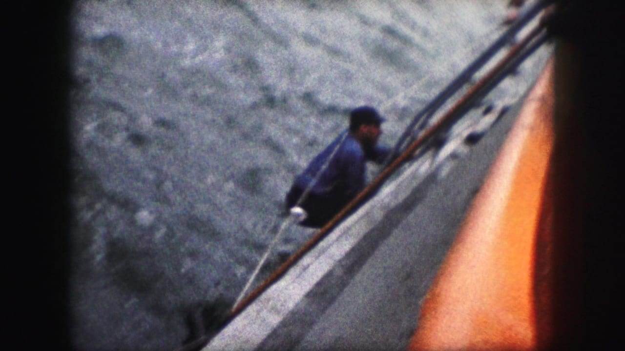 1959 8mm frame with a man in a blue shirt and dark cap beside the rail of a vessel, with choppy water below.