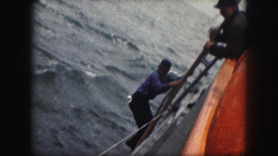 1959 8mm frame showing a man in blue gripping the vessel rail near the orange-red side, with rough water in the background.