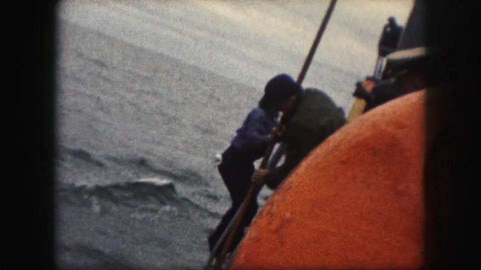 1959 8mm frame showing a person in blue near the orange-red vessel side, with another figure by the railing and open water beyond.