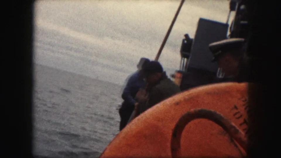 1959 8mm frame showing sailors on a ship deck near the rail, with an orange life preserver in the foreground and open water beyond.