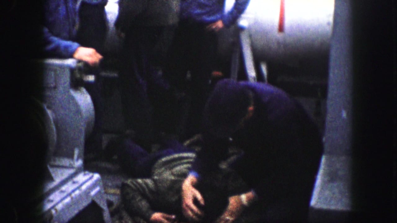 1959 Kansas City home-movie frame showing uniformed men around a person on the floor in a confined industrial space.