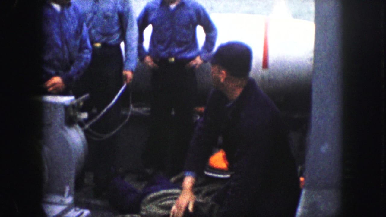 A man in a dark cap kneels beside a person on the floor while uniformed men stand nearby.