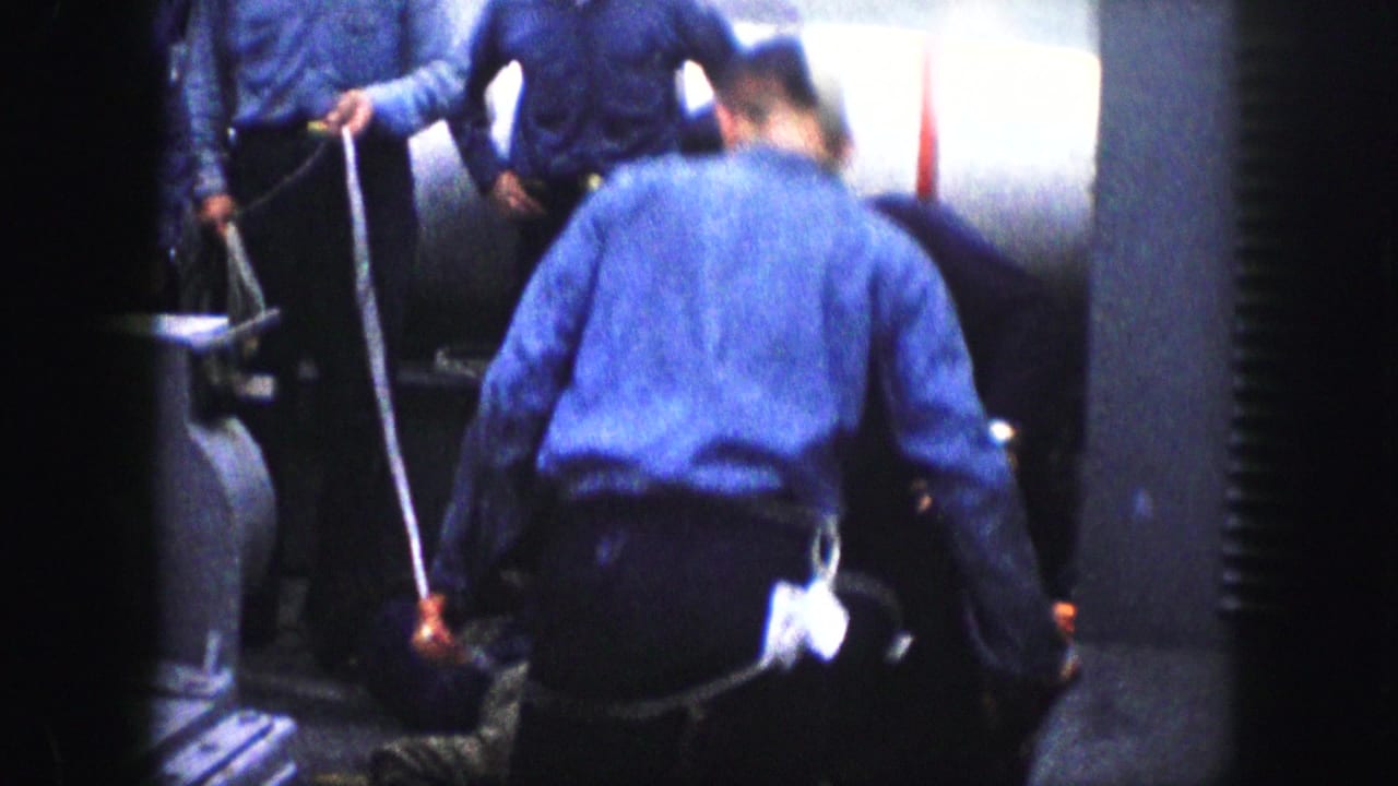 A blue-uniformed man bends into the foreground as others stand around a person down on the floor in the 1959 home-movie scene.