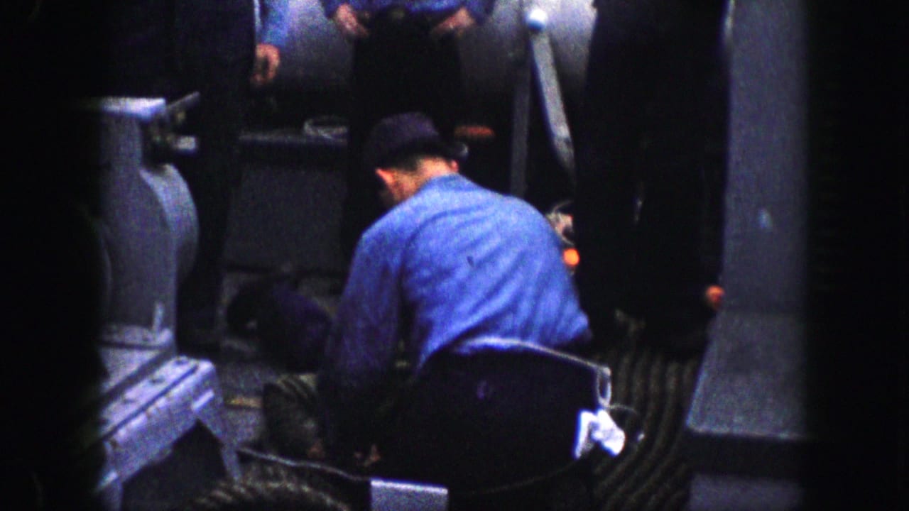 In a confined industrial or vessel-like area, a uniformed man leans over the person on the floor while another man stands nearby.