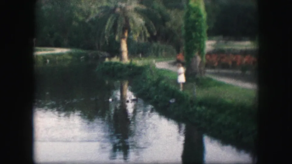 A person stands near a Kansas City park lake, with trees, plantings, and reflections visible in the 1959 8mm frame.
