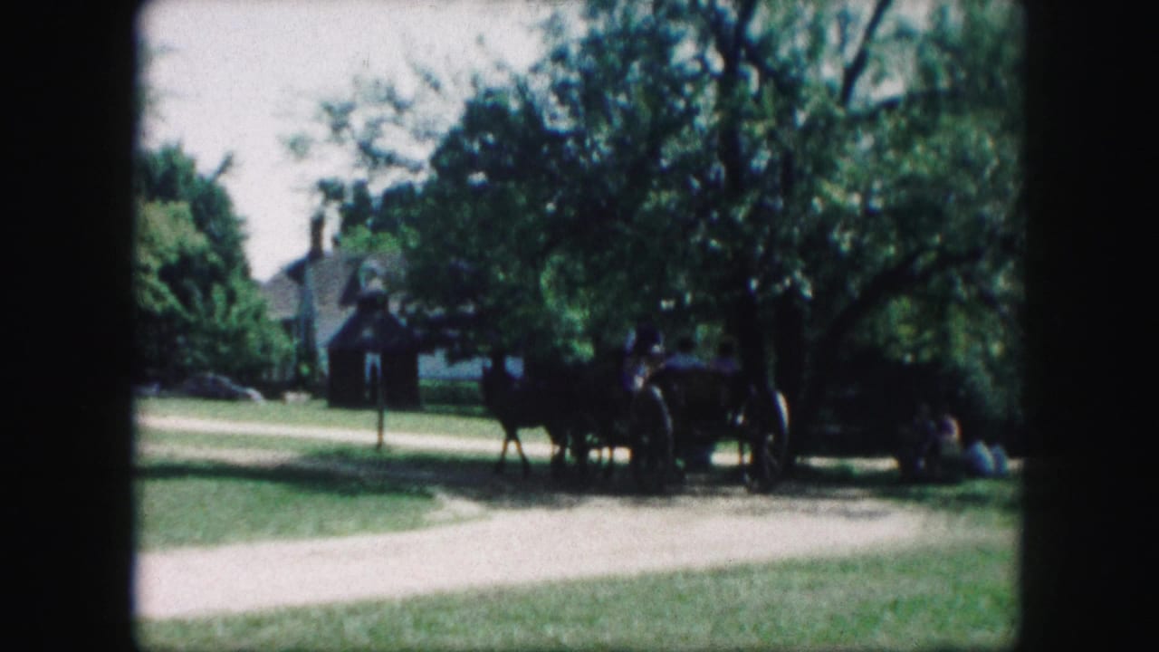 Horse team and wagon passing across grass near trees and a small building.