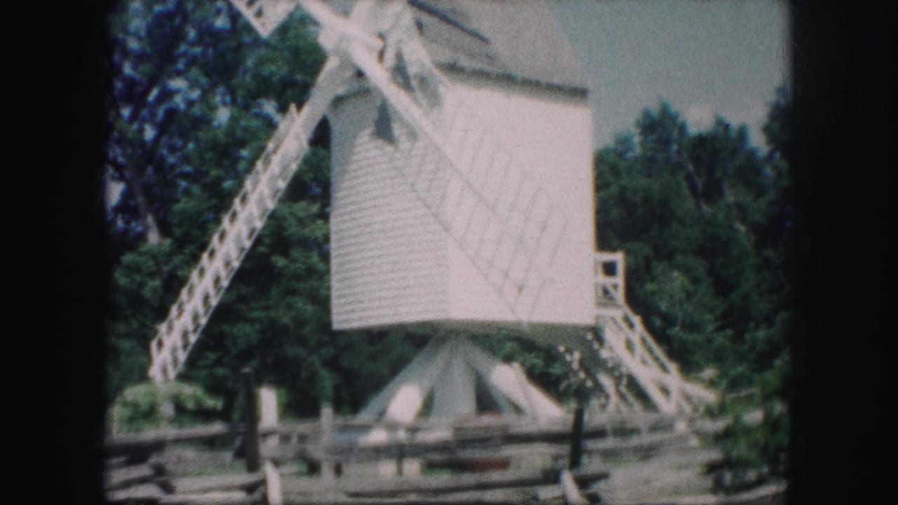 1959 Kansas City home-movie frame showing a small white windmill with lattice blades, trees, pale sky, and a split-rail fence.
