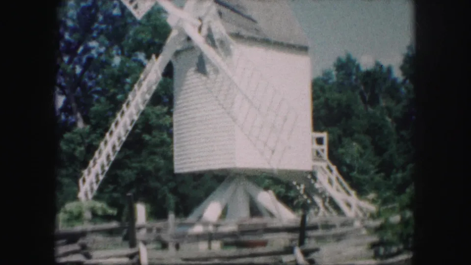 1959 Kansas City home-movie frame showing a small white windmill with lattice blades, trees, pale sky, and a split-rail fence.