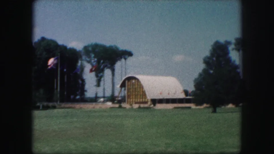 1959 Kansas City 8mm frame showing an arched white-roofed building beyond grass, with flags and trees nearby.