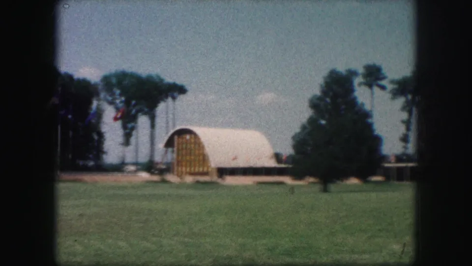 Kansas City 1959 home-movie frame of an arched barn-like building across a lawn, with flags and trees behind it.
