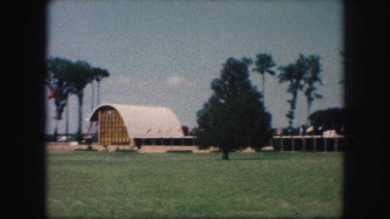 1959 Kansas City home-movie frame showing a white arched exhibition pavilion beyond a lawn, with trees and small flags nearby.