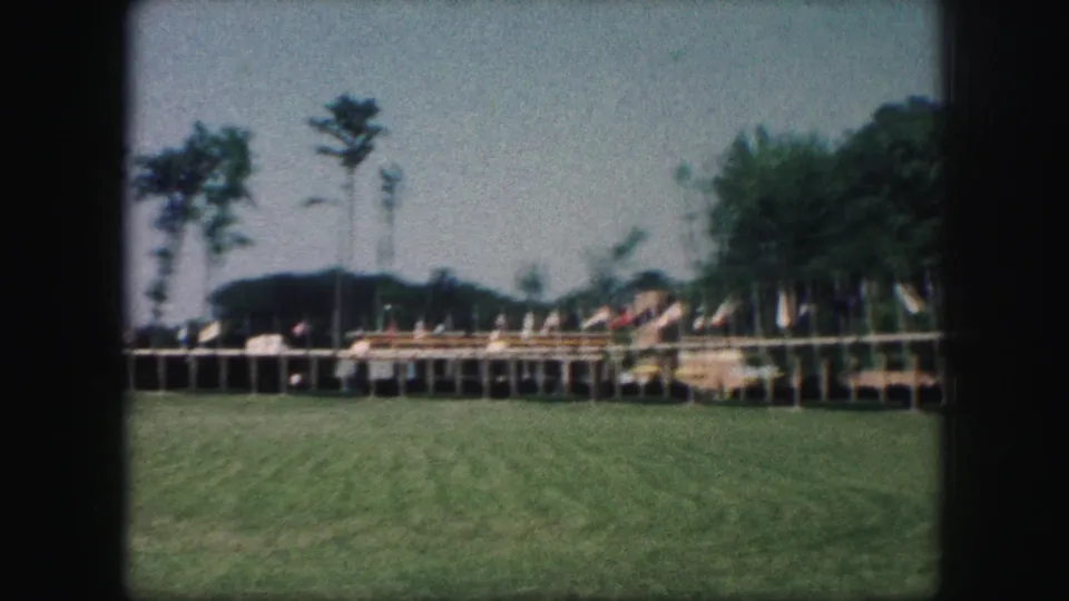 1959 Kansas City frame showing the flag-lined exhibition platform, grassy foreground, trees, and indistinct visitors in the distance.