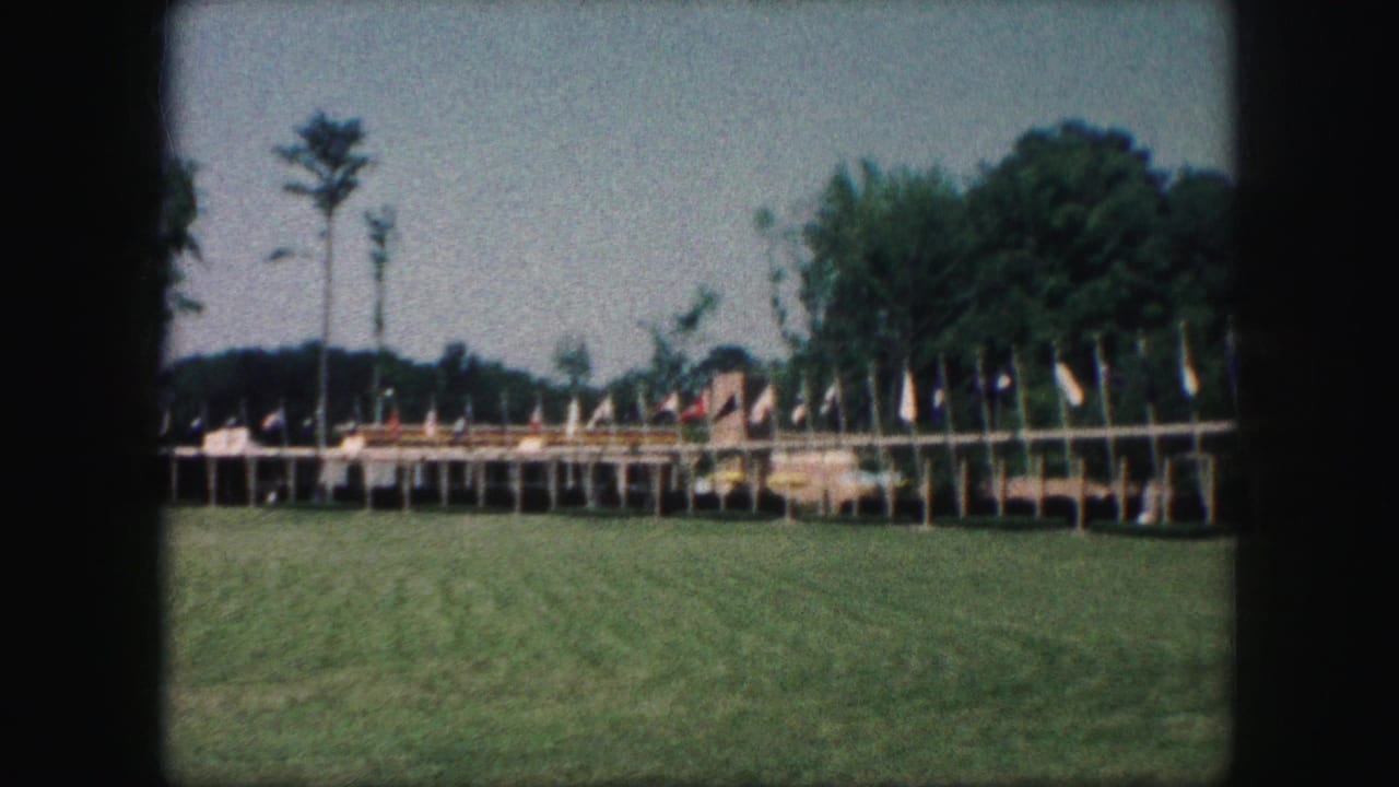 1959 Kansas City 8mm frame showing a lawn, many flags on poles, low display structures, and trees in the background.