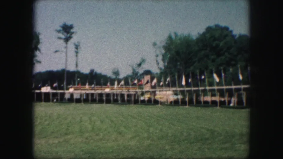 1959 Kansas City 8mm frame showing a lawn, many flags on poles, low display structures, and trees in the background.