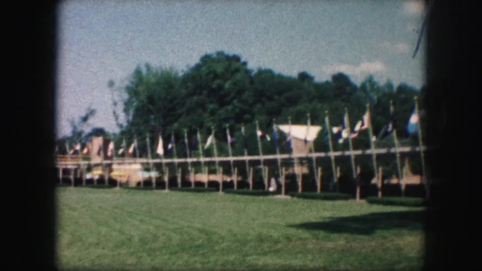 A mid-century home-movie view shows clustered flags on poles beside low structures, with a green field and trees visible.