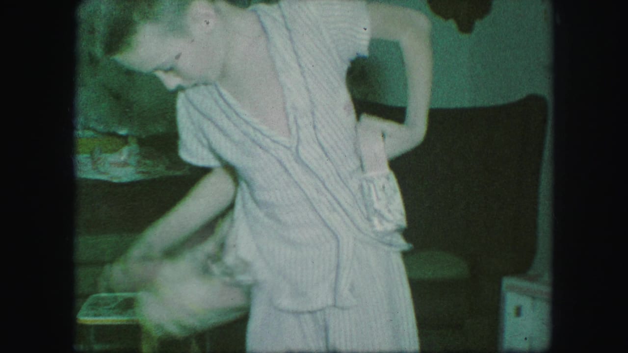 In supported 1959 Kansas City home-movie footage, a child in striped pajamas bends while handling a cowboy belt or holster indoors.