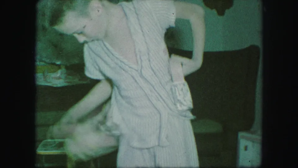 In supported 1959 Kansas City home-movie footage, a child in striped pajamas bends while handling a cowboy belt or holster indoors.