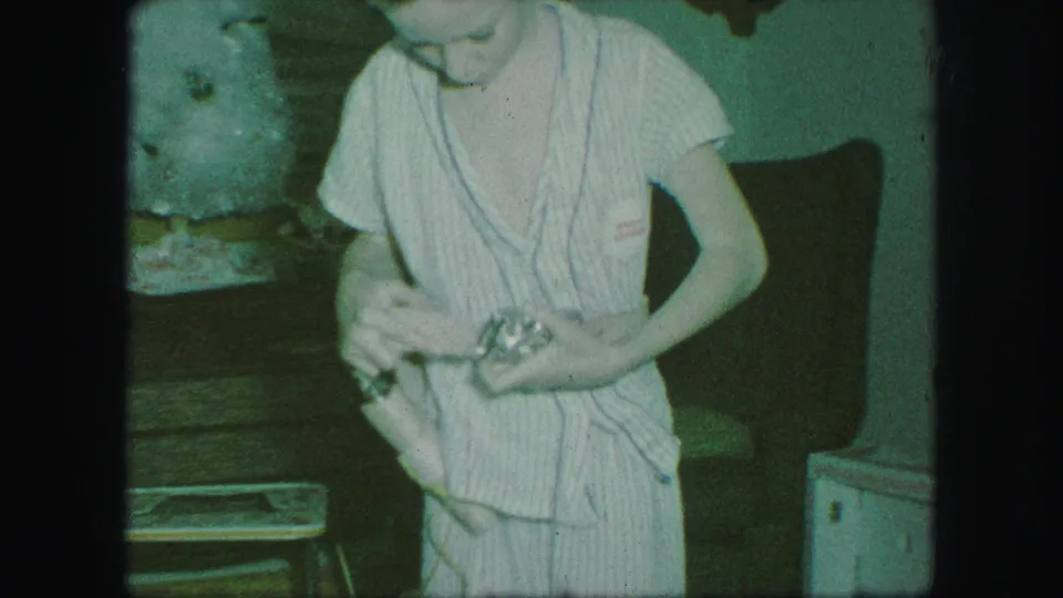 A child in striped pajamas stands in a private interior, holding and adjusting a cowboy belt or holster in 1959 home-movie footage.