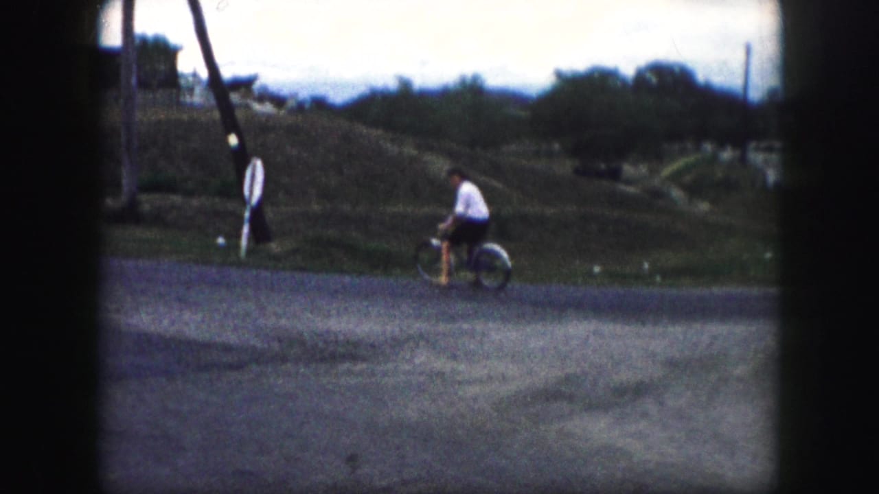 In 1959 Kansas City home-movie footage, a woman rides a bicycle across a quiet residential street near open lots and utility poles.