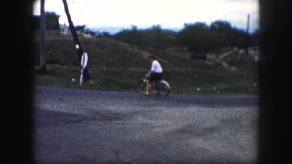 In 1959 Kansas City home-movie footage, a woman rides a bicycle across a quiet residential street near open lots and utility poles.