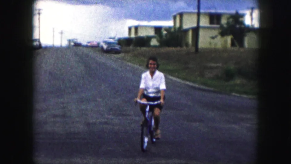 The rider smiles while bicycling toward the camera in 1959 Kansas City footage, with residential buildings and cars visible up the road.