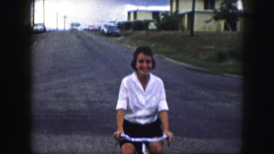 A closer 1959 Kansas City frame shows the woman smiling on her bicycle, with the residential street receding behind her.