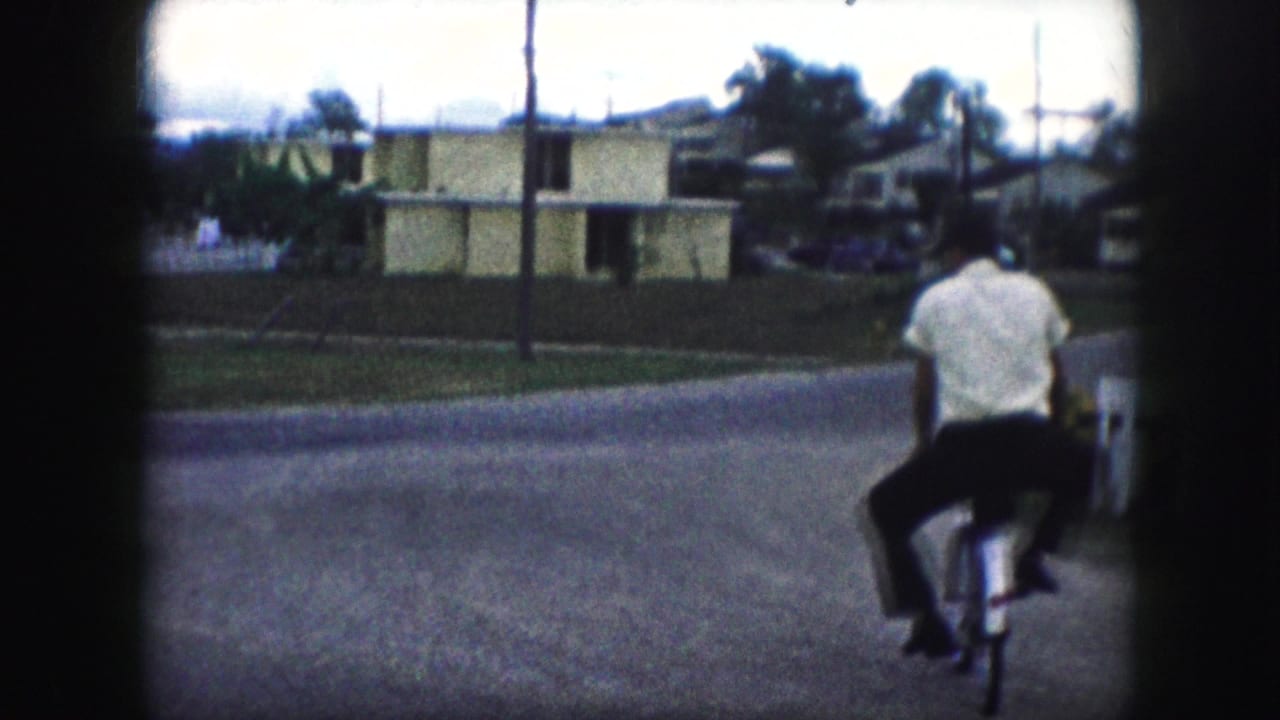A young man in a white shirt rides a bicycle along a residential street in the 1959 Kansas City home-movie scene.