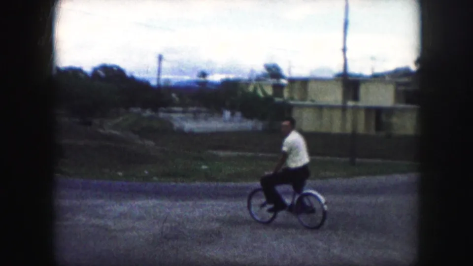 The bicyclist moves across the frame with low houses, lawns, and utility poles visible in the Kansas City street setting.