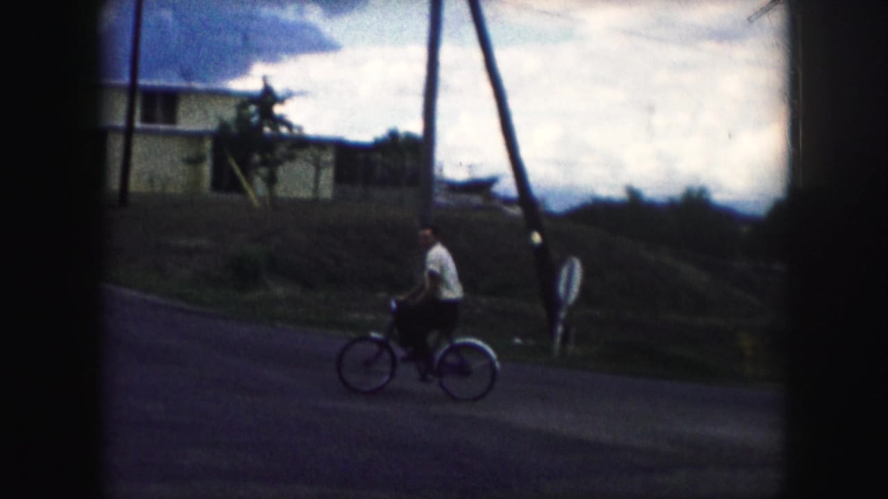 In 1959 Kansas City home-movie footage, a man in a light shirt pedals a bicycle across a quiet residential street.