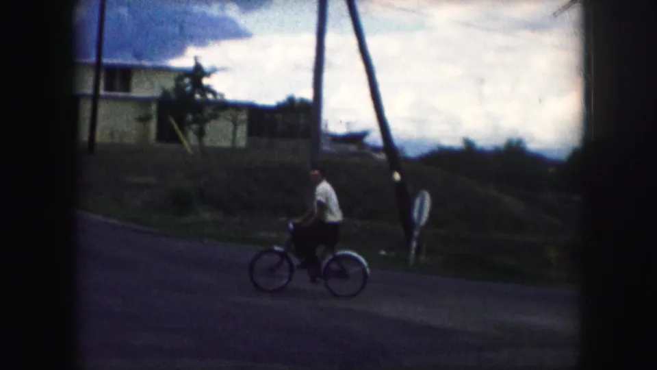 In 1959 Kansas City home-movie footage, a man in a light shirt pedals a bicycle across a quiet residential street.