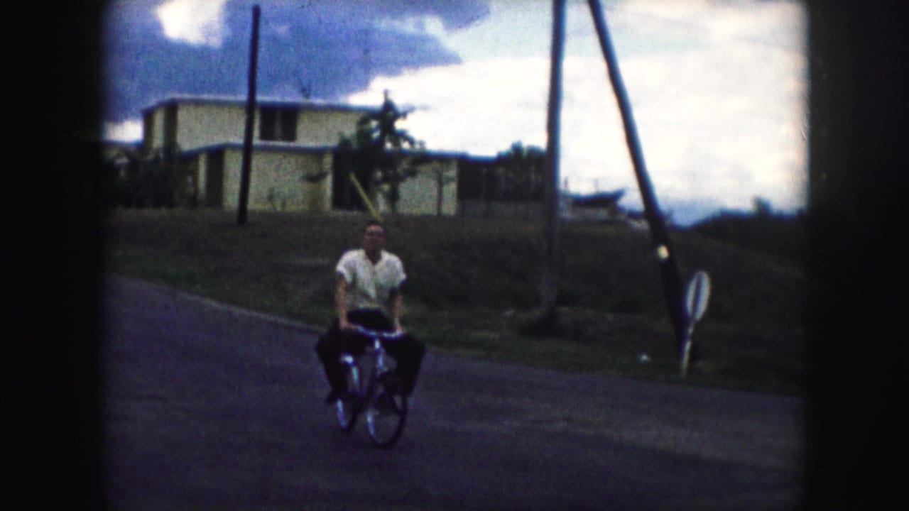 The cyclist turns toward the camera on a Kansas City residential street, with houses and utility poles behind him.