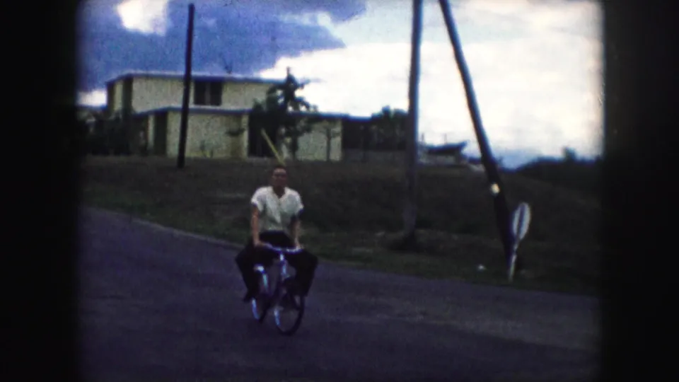 The cyclist turns toward the camera on a Kansas City residential street, with houses and utility poles behind him.