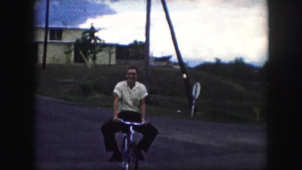 A smiling adult man in a light short-sleeved shirt rides closer to the camera in 1959 Kansas City footage.