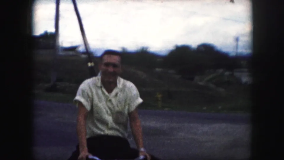 Close home-movie frame of the bicyclist smiling as he rides toward the camera on a Kansas City residential street.