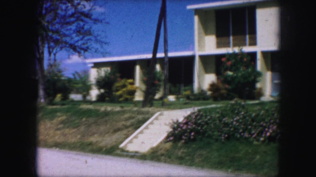 1959 Kansas City home-movie frame showing a white two-story modern building with broad windows, lawn, walkway, and flowering landscaping.