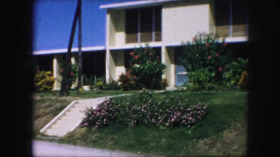 1959 Kansas City 8mm frame with the modern building facade closer in view, showing windows, shaded entrance area, lawn, and flowering shrubs.