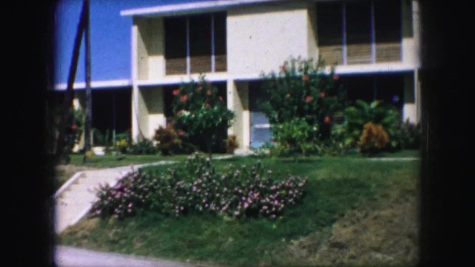 1959 Kansas City home-movie frame showing the white modern exterior, green lawn, flower beds, walkway, and dark window bays.
