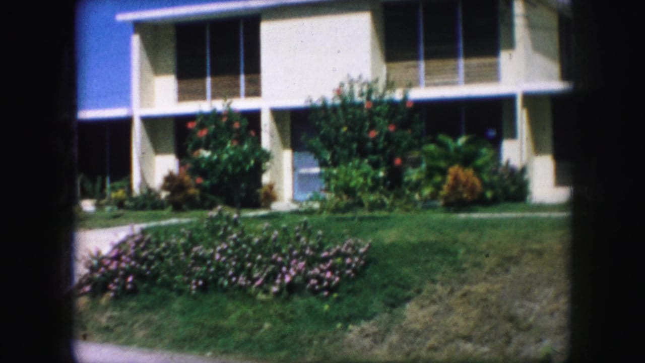 1959 Kansas City home-movie frame showing a cream two-story house with large windows, a covered entry, shrubs, lawn, and pink flowers.