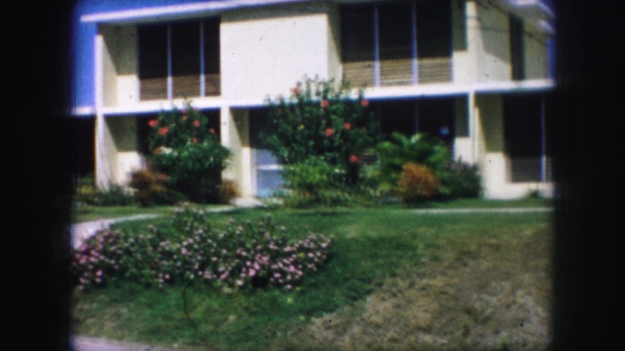 1959 Kansas City residential exterior with a manicured lawn, pink flower bed, shrubs, and a cream house facade in soft 8mm color.