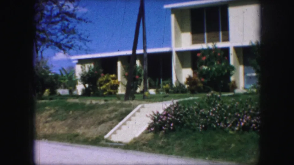 Wider 1959 Kansas City residential frame showing the cream house, concrete walk, flower bed, shrubs, lawn, blue sky, and a tree trunk.