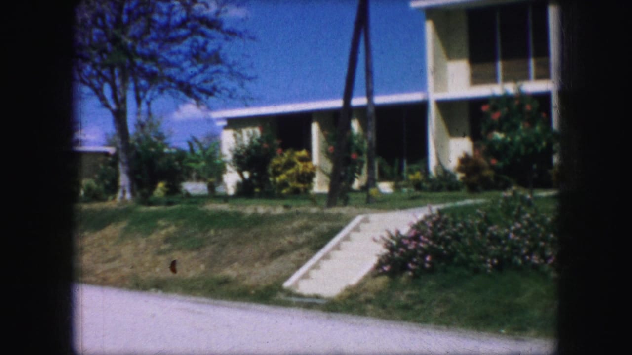 1959 Kansas City home-movie frame showing a pale suburban house, sloped lawn, concrete ramp, flowering shrubs, tree, utility poles, and road.