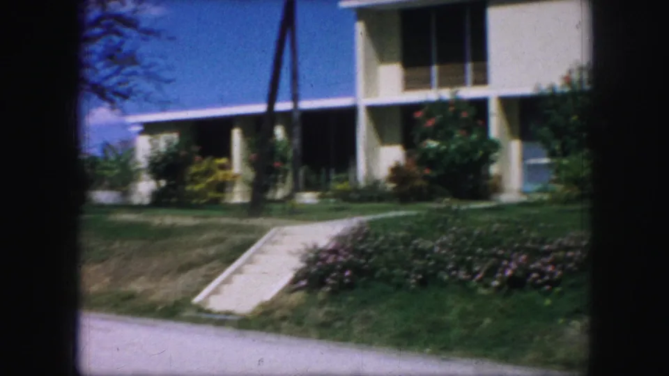 Kansas City 1959 home-movie frame with a closer view of the pale house, green lawn, concrete ramp, flowering bushes, roadside, and pole shadows.