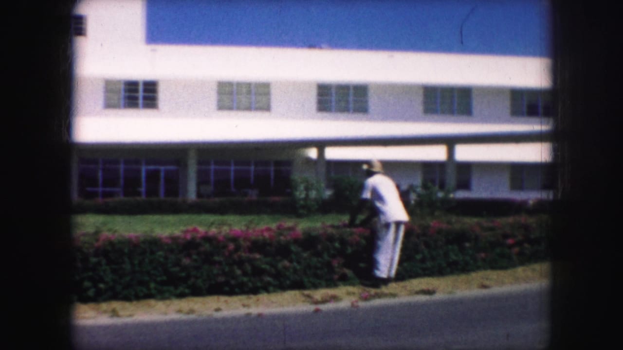 Gardener bends near hedges in front of a white multi-story building.