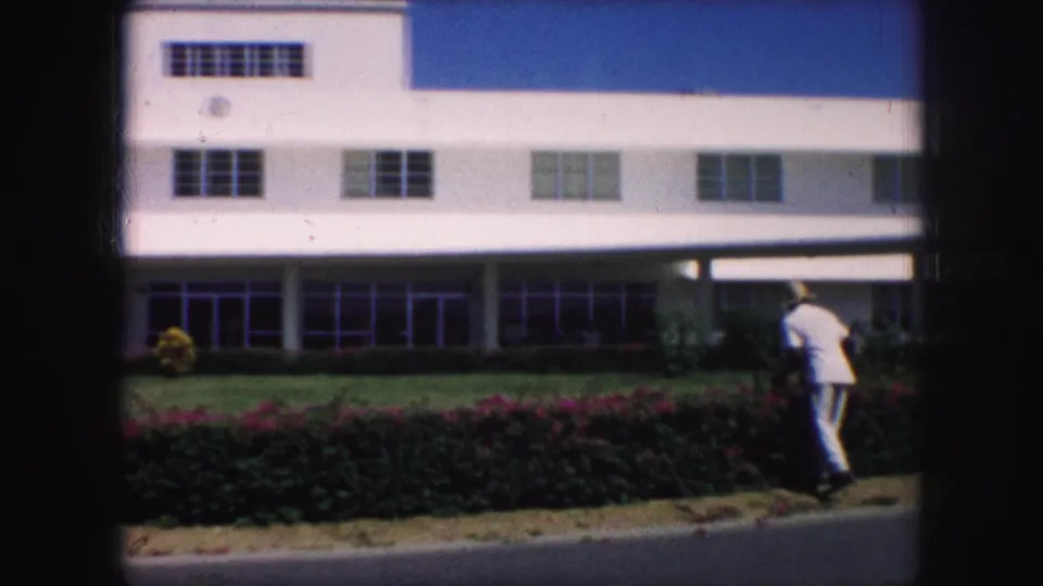 Kansas City 1959 home-movie view of a gardener pruning hedges along a road before a white building facade.