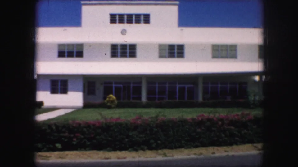 Kansas City 1959 frame showing the white building front, lawn, hedges, walkway, and clear sky without the gardener in view.