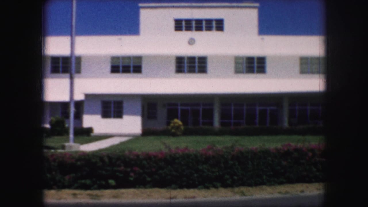 1959 Kansas City home-movie frame showing a white mid-century building facade with rows of windows, lawn, hedges, sidewalk, and flagpole.