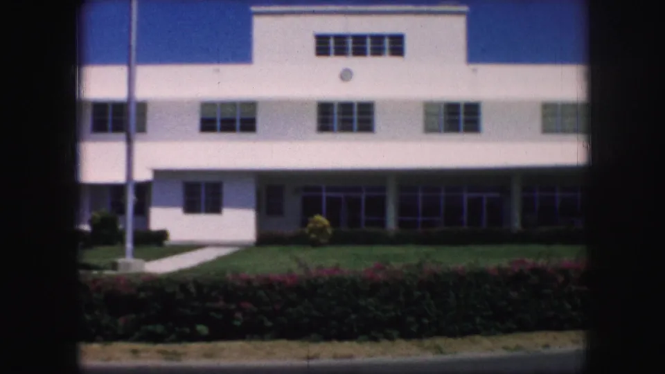 1959 Kansas City home-movie frame showing a white mid-century building facade with rows of windows, lawn, hedges, sidewalk, and flagpole.