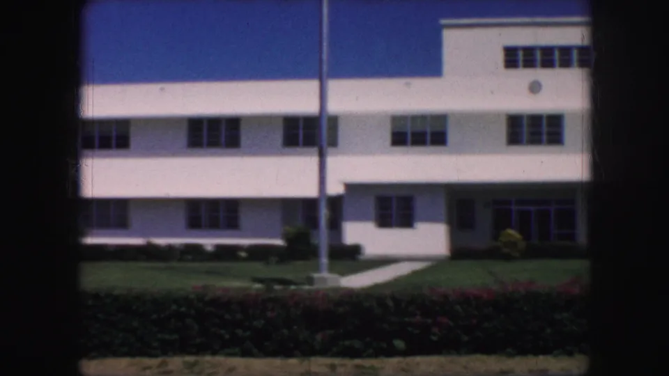 1959 Kansas City home-movie frame with the white building cropped closer, showing window bands, flagpole, sidewalk, lawn, and hedges.