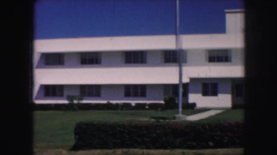 1959 Kansas City frame showing a partly cropped white mid-century facade with rows of windows, flagpole, lawn, hedges, and clear sky.