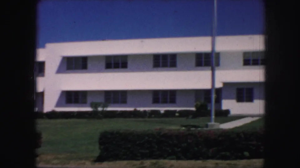 A 1959 Kansas City exterior shows a white multi-story building, flagpole, lawn, and hedge in soft 8mm color.